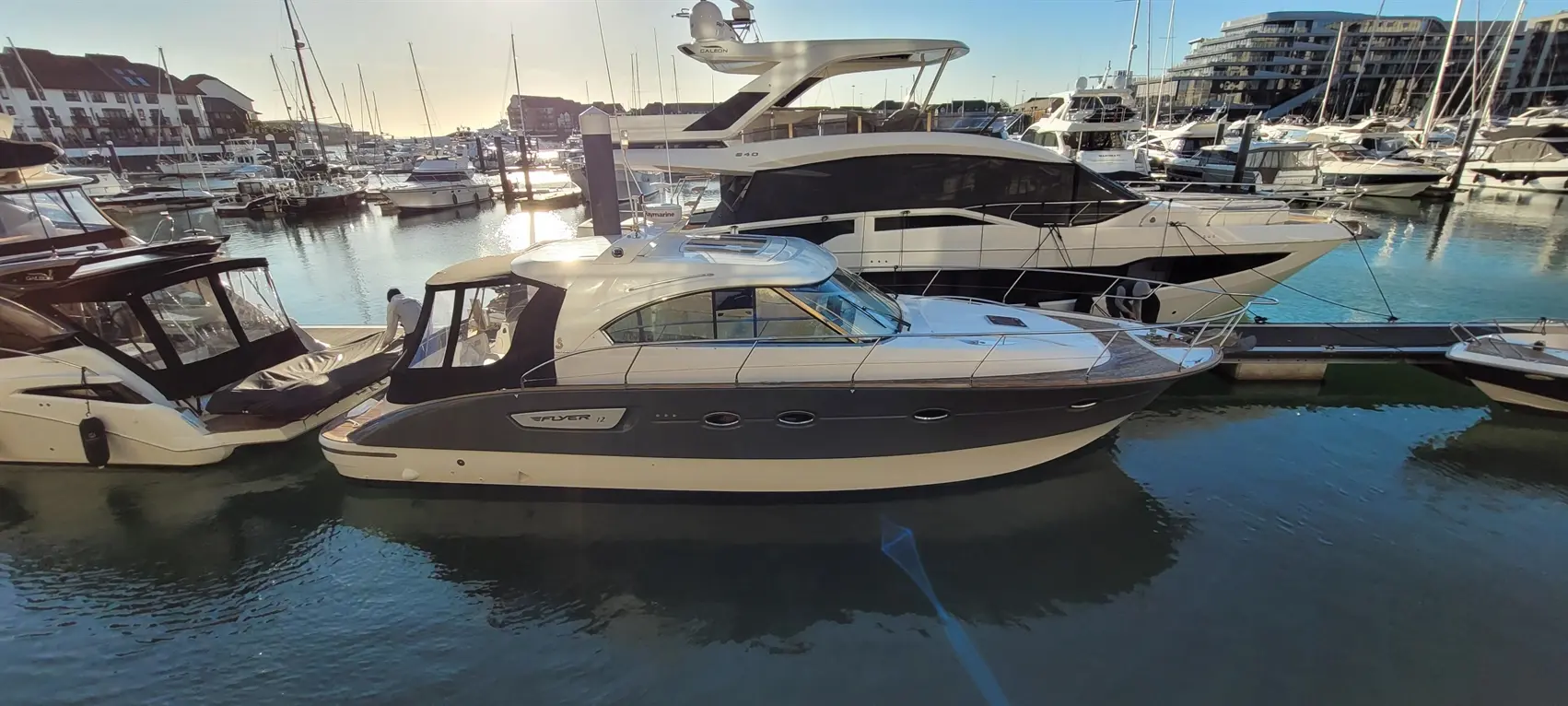 A sleek Flyer 12 motorboat docked at a marina, with additional boats and waterfront buildings in the background, reflecting sunlight.